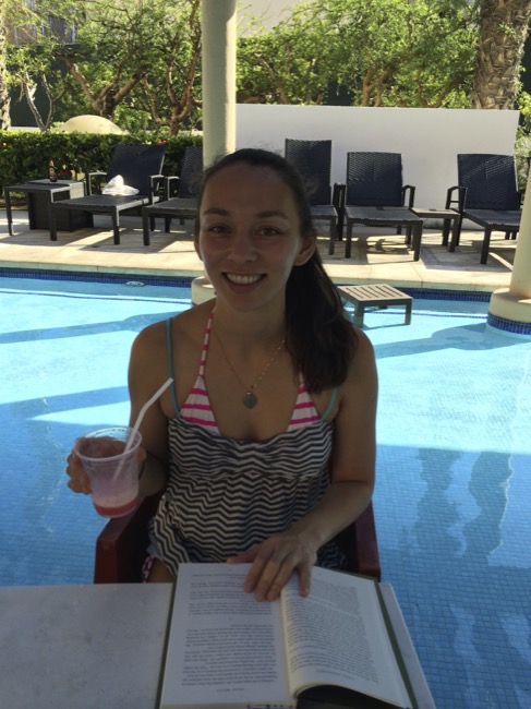 a woman sitting in a chair by a pool holding a drink and a book