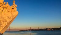 a statue of a man and woman on a cliff above water with a bridge in the background