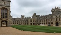 a large stone building with a lawn and cars parked in front of it with Windsor Castle in the background