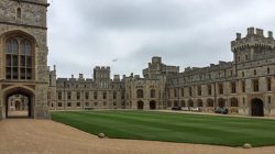 a large stone building with a lawn and cars parked in front of it with Windsor Castle in the background