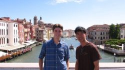 two men standing on a bridge over a river with buildings in the background