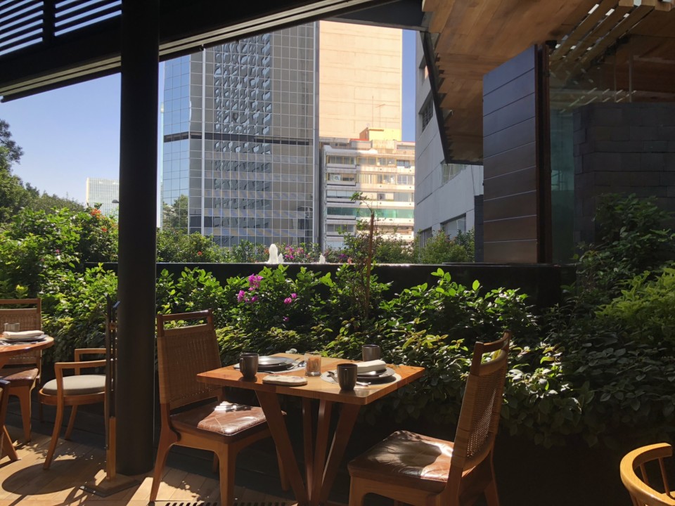 a table and chairs outside with plants and buildings in the background