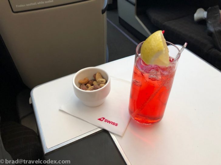a glass of red liquid with a lemon wedge and a bowl of nuts