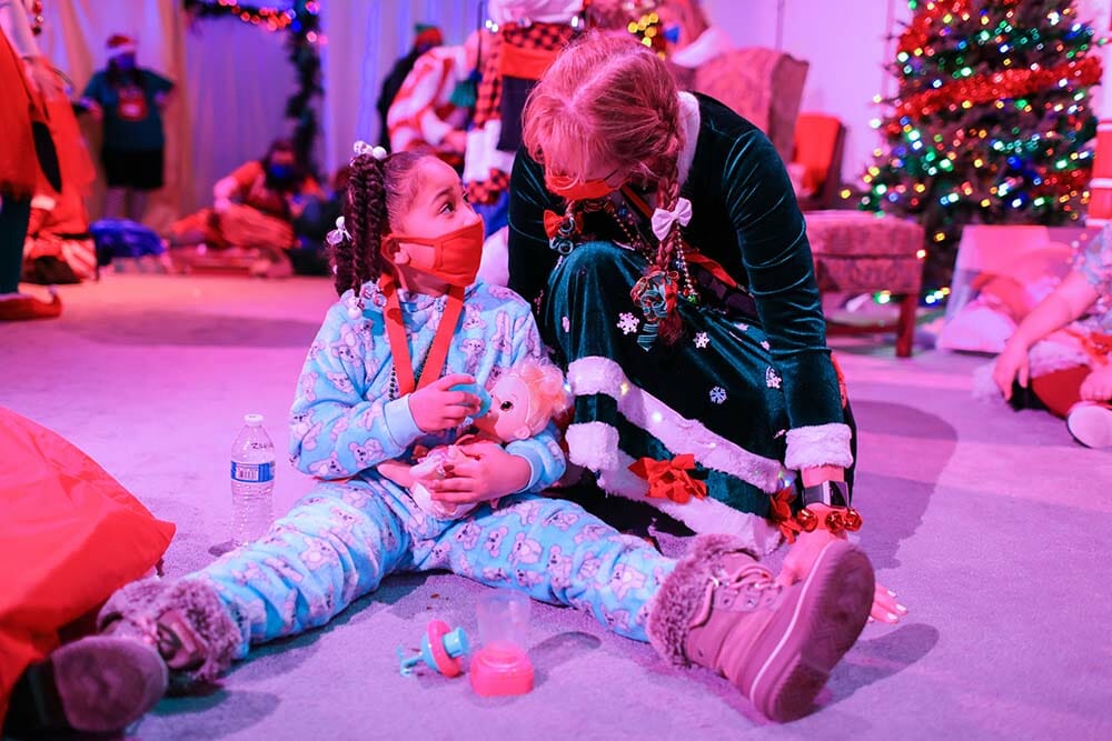 a girl sitting on the floor with a doll and a woman wearing a mask